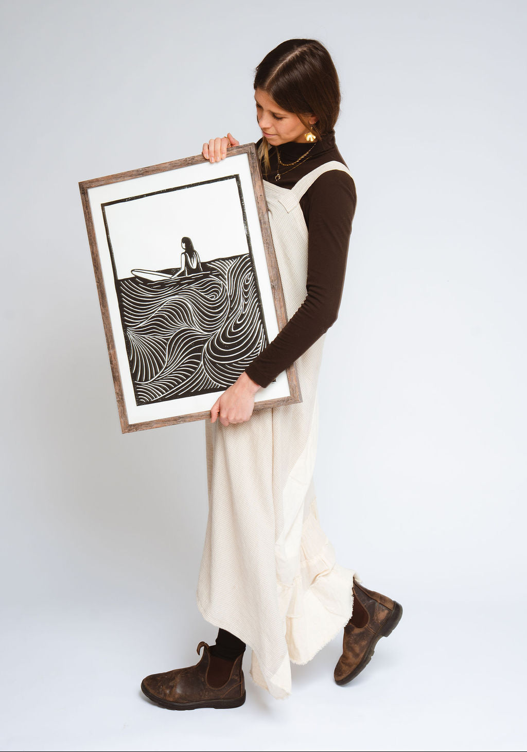 Emily Wright artist in a cream dress and brown boots holding her framed black and white art print featuring a minimalist figure on a wavy line pattern, set against a light gray studio background.