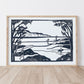 Framed black and white artwork of a lino print coastal scene with people and surfboards on a white wall.