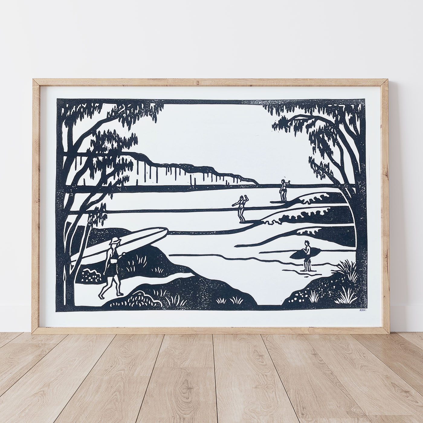 Framed black and white artwork of a lino print coastal scene with people and surfboards on a white wall.
