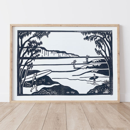 Framed black and white artwork of a lino print coastal scene with people and surfboards on a white wall.