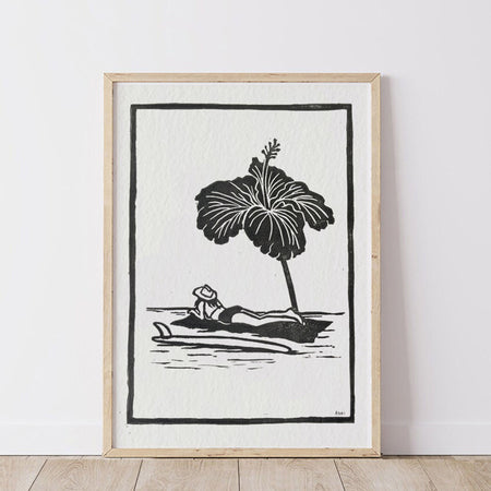 Framed black and white artwork of a woman lounging under a hibiscus flower as a beach umbrella.