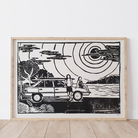 Framed black and white artwork of two people by a vintage car with surfboards on the roof with a scenic coastal background.