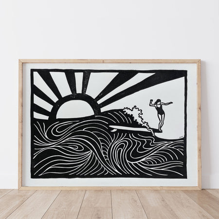 Framed artwork of a surfer nose riding on a wave with a sun in the background on a white wall.