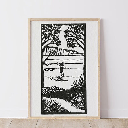 Framed black and white  lino print artwork of landscape with a person surfing in nature with trees, waves  and cliffs