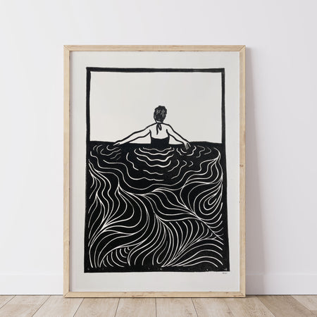 Framed black and white lino print artwork of a person swimming / wading in water on a white wall.